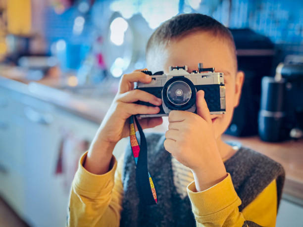 A young boy in a yellow and gray sweater holds a toy camera designed to look like a vintage model, standing in a brightly lit kitchen with a blurred background.