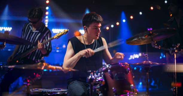 istock Handsome Young Drummer Performing on Concert Stage with His Band Members, Surrounded by Falling Confetti and Pulsating Visuals. Young Musicians Showcasing Their Skills and Energy. 2185403925
