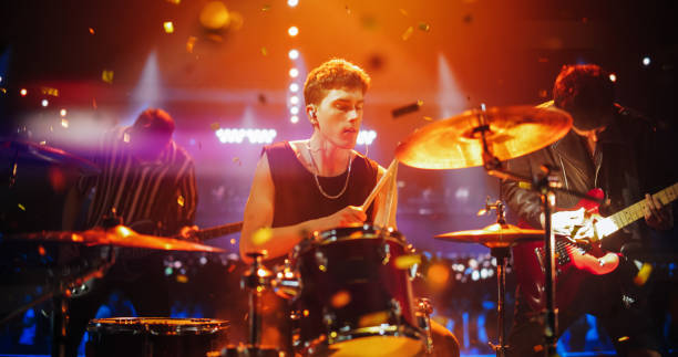 istock Handsome Young Drummer Performing on Concert Stage with His Band Members, Surrounded by Falling Confetti and Pulsating Visuals. Young Musicians Showcasing Their Skills and Energy. 2185403961