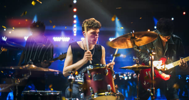 Handsome Young Drummer Performing on Concert Stage with His Band Members, Surrounded by Falling Confetti and Pulsating Visuals. Young Musicians Showcasing Their Skills and Energy. Slow Motion Footage