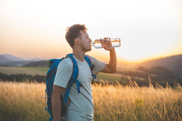 istock Hiker with a blue backpack drinks from a water bottle while standing in a sunlit field at sunset. The golden light, distant hills, and peaceful surroundings emphasize the connection with nature 2185416694