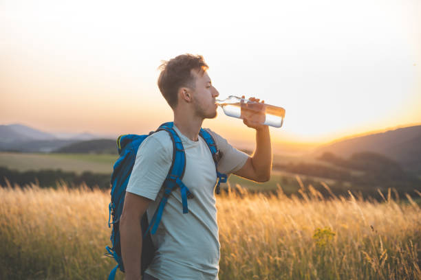 istock Hiker with a blue backpack drinks from a water bottle while standing in a sunlit field at sunset. The golden light, distant hills, and peaceful surroundings emphasize the connection with nature 2185416696
