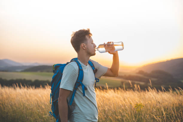istock Hiker with a blue backpack drinks from a water bottle while standing in a sunlit field at sunset. The golden light, distant hills, and peaceful surroundings emphasize the connection with nature 2185416699