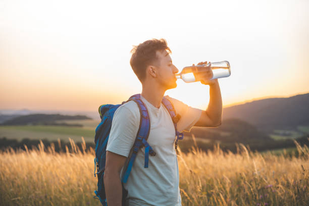 Hiker with a blue backpack drinks from a water bottle while standing in a sunlit field at sunset. The golden light, distant hills, and peaceful surroundings emphasize the connection with nature