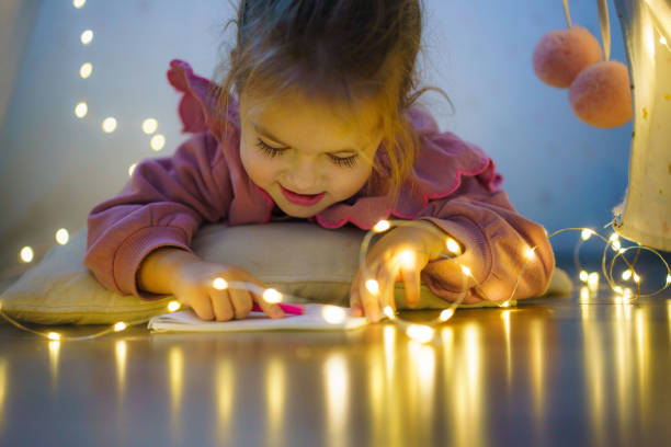 Happy little girl lying down and drawing whit crayon in the illuminated tent