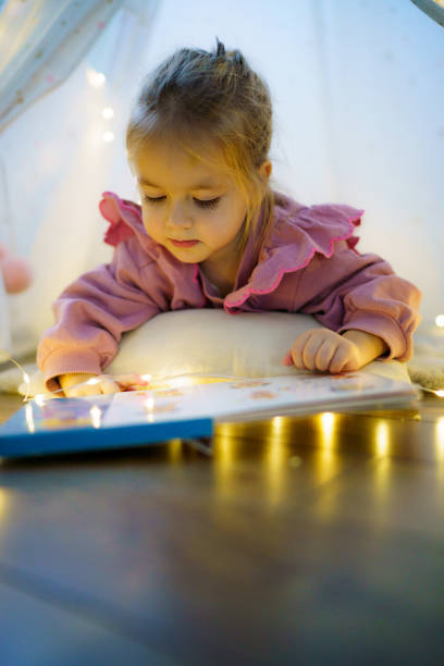 istock Cute girl looking at picture book while lying in tent 2185425298