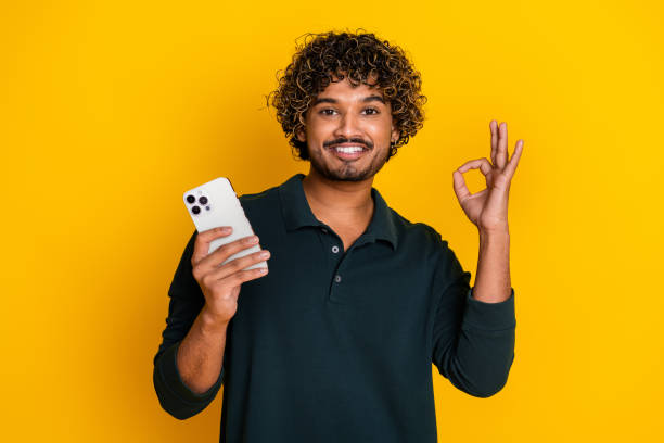 istock Confident young man makes OK gesture holding smartphone against vibrant yellow background 2185432975