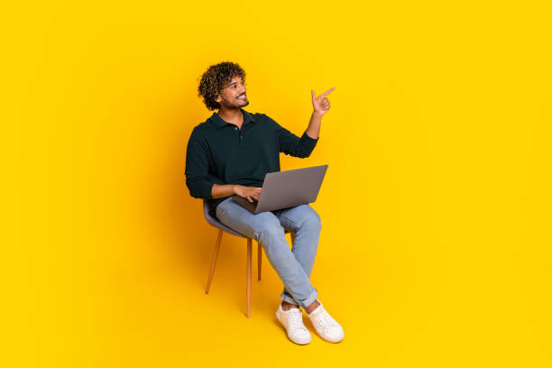 istock Young man in casual attire using laptop while pointing up with curiosity on vibrant yellow background. 2185433083