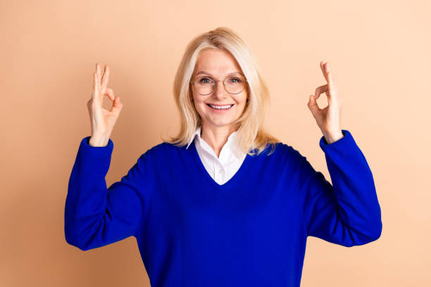 istock Confident mature businesswoman gesturing okay with smile and style in blue pullover against beige background 2185444505