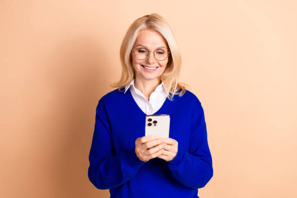 istock Mature woman in blue pullover holding smartphone against beige background, smiling confidently 2185444603