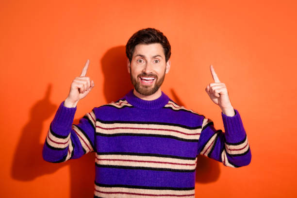 istock Cheerful man in striped sweater points up against vibrant orange backdrop, radiating positivity 2185444708