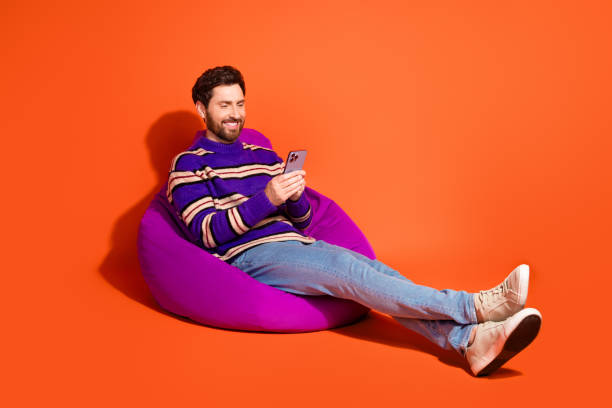 istock Confident man relaxing on a bean bag while using smartphone, vibrant orange background and stylish outfit create a cheerful atmosphere 2185444825
