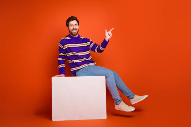 istock Full body photo of attractive young man sit cube point empty space dressed stylish striped purple clothes isolated on orange background 2185445171