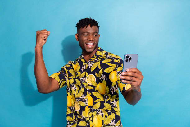 istock Joyful young man celebrating success while using phone against blue background in stylish summer shirt 2185445488