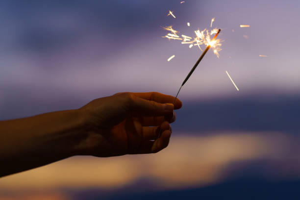 istock Woman's hand holding a bright sparkler on a blue sky background with sunset. Concept of celebration, New Year greetings, holidays, miracles, Christmas 2185463674