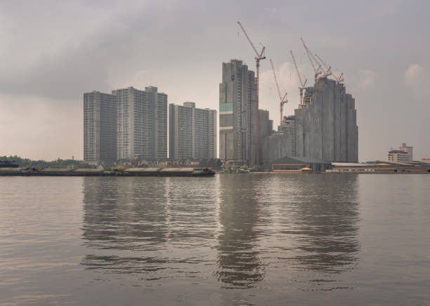 Bangkok, Thailand - 17 Nov, 2024 - Skyscrapers and Residential buildings complex of under construction long the chao phraya river at afternoon. View of Modern high-rise buildings against sky background with reflection in the surface of water, Tower cranes in action, use it as your wallpaper, poster and Space for text, Selective focus.