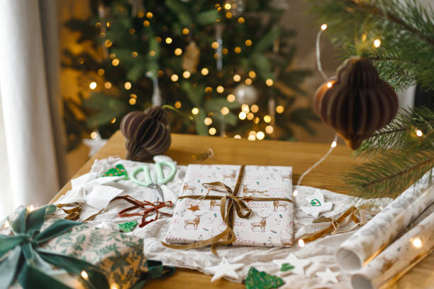 istock Merry Christmas! Stylish christmas gift, modern paper ornaments and pine branches on table against golden christmas tree lights. Atmospheric christmas eve. Modern wrapped present 2185489213