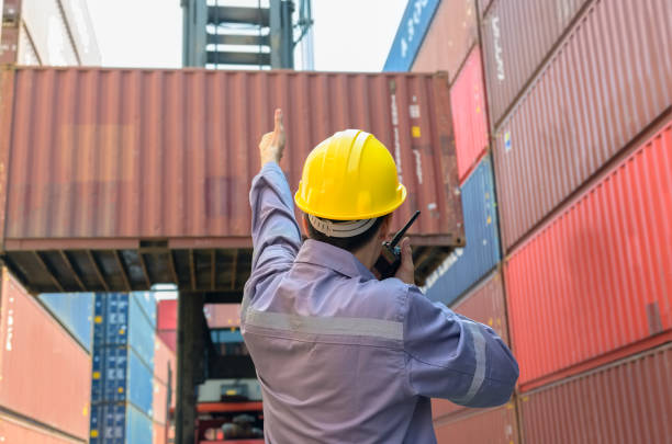 istock Dock supervisor in yellow hard hat and uniform directing container loading with walkie talkie and thumb up gesture at shipping container yard. 2185509249