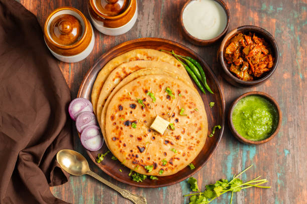 Aloo paratha, a golden-brown Indian flatbread stuffed with spiced mashed potatoes, is served with tangy achar (pickle) and refreshing chutney, creating a perfect blend of flavors for a comforting, savory delight.