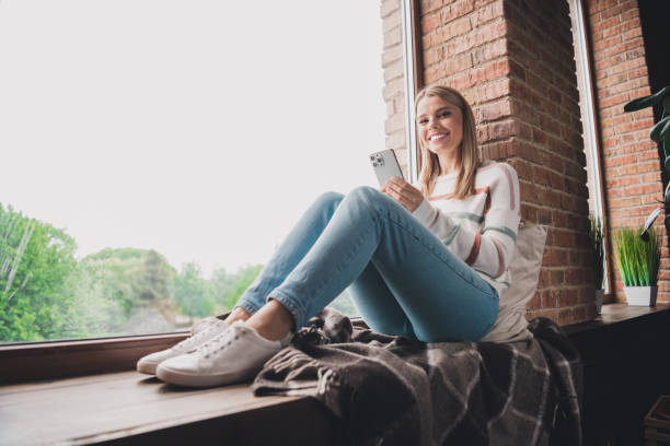 istock Young woman enjoying a relaxing afternoon by the window, smiling while using her smartphone in the comfort of her home 2185513973