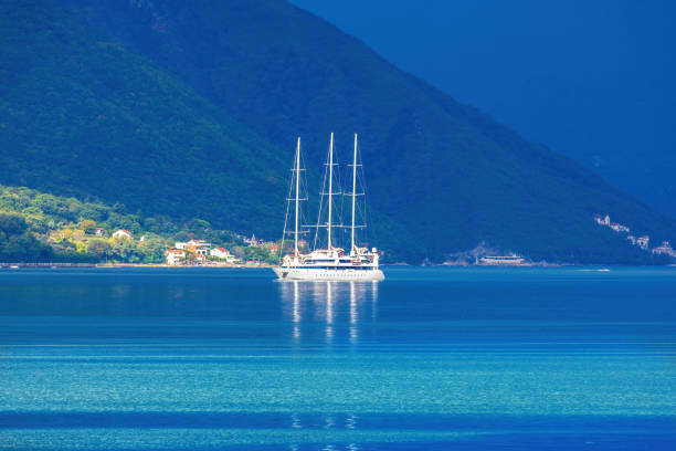 A tranquil scene in the Bay of Kotor, Montenegro, just before rainfall. A white sailboat floats on calm, turquoise waters, framed by lush green mountains under a deep blue sky.