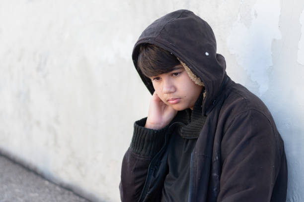 Unhappy young person lowering head, sitting alone on pavement against white brick wall, portraying sadness and loneliness