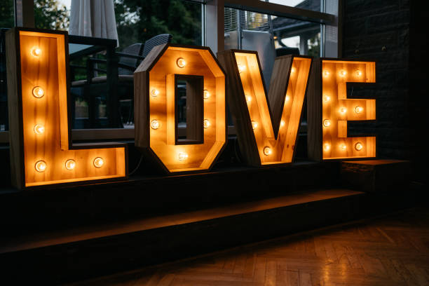 Large wooden marquee LOVE letters illuminated with warm light bulbs displayed on a dark platform against window background. The decorative light installation features rustic wooden construction with exposed bulbs creating a romantic atmosphere at an indoor wedding venue. The letters cast a warm golden glow on the herringbone wooden floor, creating an intimate ambiance perfect for wedding celebrations and romantic events.