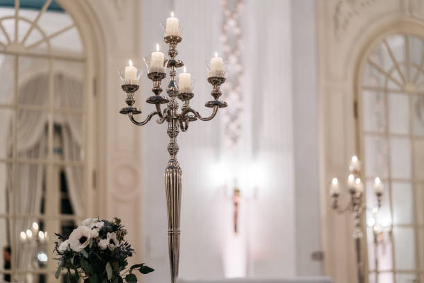 Luxurious silver candelabra featuring five white pillar candles in glass holders, set against a bright white interior with arched windows. The classic ornate candlestick holder displays intricate metalwork detailing and is accompanied by a romantic floral arrangement of white anemones and dark green foliage. The soft, warm candlelight creates an intimate atmosphere perfect for a winter wedding celebration, while architectural elements like Palladian windows add grandeur to the elegant setting.