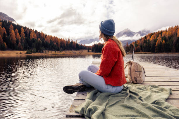 A woman in a blue beanie and orange sweater sits on a wooden dock, enjoying the tranquil view of a mountain lake surrounded by autumn trees under a cloudy sky.