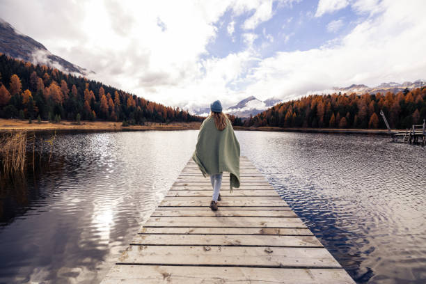 A woman wrapped in a green blanket and wearing a blue beanie walks along a wooden dock, surrounded by a serene lake and autumn-colored trees under a partly cloudy sky.