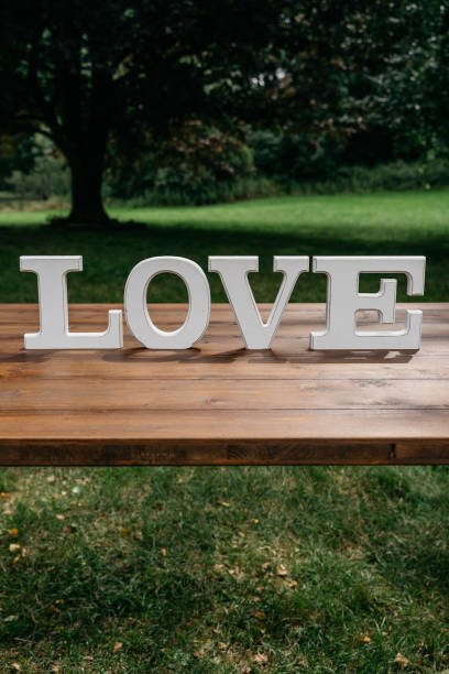 White wooden LOVE letters displayed on rustic wooden table outdoors in garden setting. Decorative freestanding wedding signage photographed in natural daylight with blurred green foliage background. Elegant minimalist wedding decor piece perfect for ceremonies and receptions.
