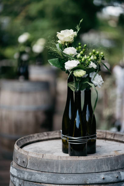 Elegant wedding decoration featuring dark wine bottles bundled together with wire, filled with fresh white roses, carnations and green berries. The minimalist floral arrangement sits atop a weathered wooden wine barrel, creating a perfect blend of rustic charm and romantic elegance. The soft bokeh background and natural lighting enhance the romantic vineyard wedding atmosphere.