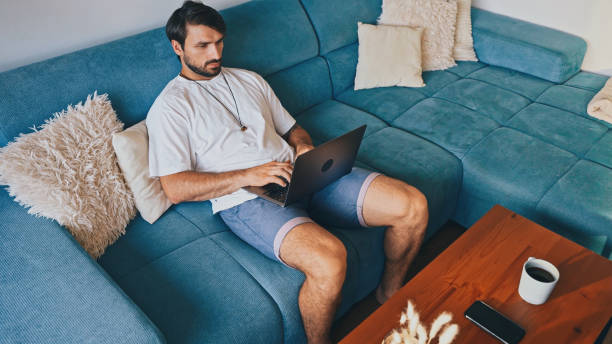 istock Relaxed Man Using Laptop on Sofa With Phone and Coffee on Table 2185585199