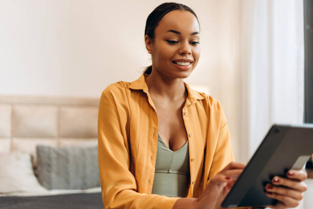 istock Happy young African American woman using tablet pc sitting on bed at home 2185587559
