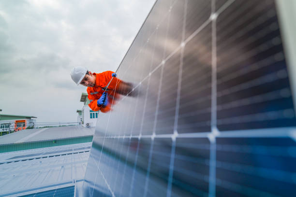 istock A worker in an orange safety jacket inspects solar panels on a rooftop, using a laptop to ensure proper maintenance. The scene highlights renewable energy, solar technology, and sustainable practices. 2185595881