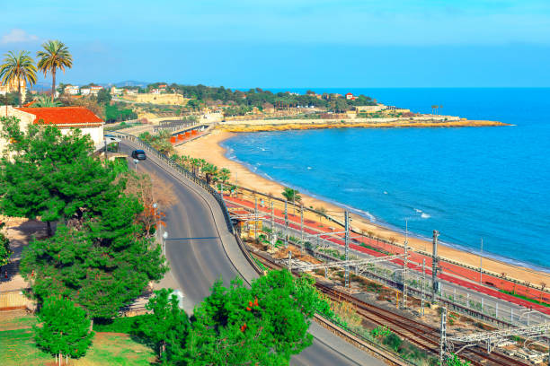 Scenic view of Tarragona, Spain, with a beautiful coastal landscape featuring a sandy beach. In the foreground, a road and railway tracks run parallel to the coastline, with an electrified rail system