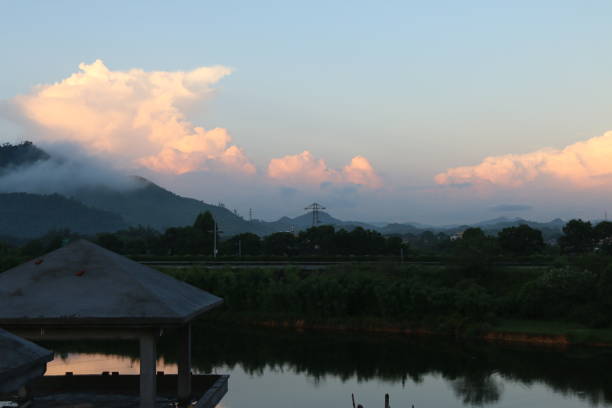 A stunning cumulonimbus cloud illuminated by the golden light of the setting sun. The cloud's warm hues contrast beautifully with the darker foreground, which features silhouettes of trees and houses in the village.