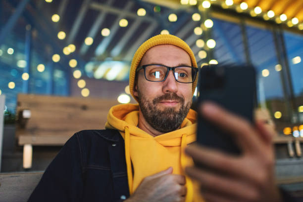 Mid adult man sitting in outdoors cafe with mobile. Cafe decorated with light bulbs.
