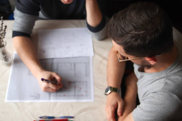 An overhead view of architects' hands working on detailed building plans. scene captures sketches and blueprints spread across a desk, with hands holding pens and rulers, actively refining designs
