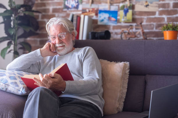 istock Smiling senior bearded man with eyeglasses relaxing on sofa at home reading a book. Retirement lifestyle concept 2185635725