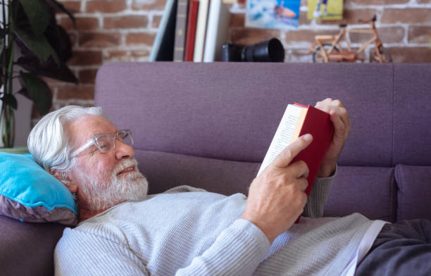 istock Smiling senior bearded man with eyeglasses relaxing on sofa at home lying down and reading a book. Retirement lifestyle concept 2185635847