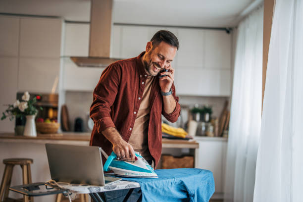 istock A mid-adult, handsome, cheerful man is ironing clothes while the kitchen is visible in the background. 2185637794