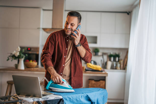 istock A mid-adult, handsome, cheerful man is ironing clothes while the kitchen is visible in the background. 2185637816