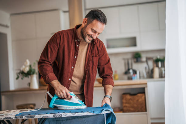 istock A mid-adult, handsome, cheerful man is ironing clothes while the kitchen is visible in the background. 2185638590