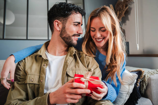 istock Joyful Young Couple Sharing Intimate Moment on Couch as Boyfriend Holds Red Gift Box, Both Radiating Happiness During Special Holiday Celebration in Their Cozy Living Room 2185654737