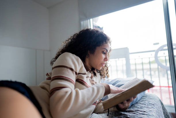 istock Young woman reading a book on bed at apartment 2185701754