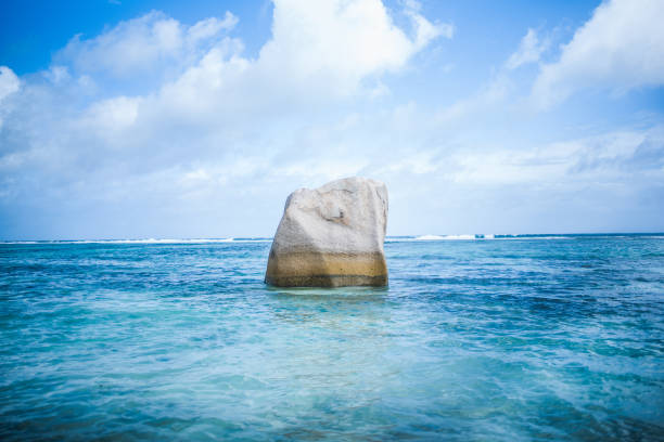 A solitary granite boulder emerges from crystal-clear turquoise waters under a vibrant blue sky with scattered clouds. The tranquil seascape exudes a sense of isolation and natural beauty.