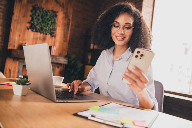 istock Confident businesswoman in modern office checking phone while working on laptop with a smile, showcasing success and multitasking skills 2185710590