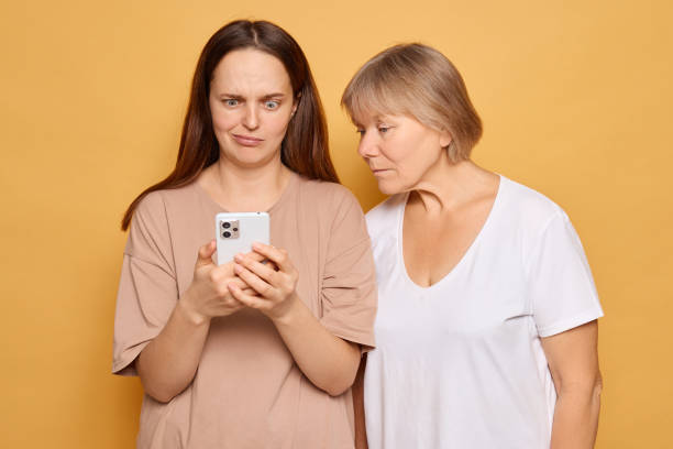 istock Two women engage with a smartphone, displaying mixed emotions in front of a warm yellow backdrop, capturing a moment of curiosity and surprise during their interaction 2185711261