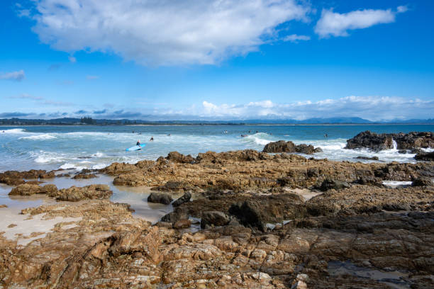 A stunning rocky beach at Byron Bay with surfers riding gentle waves under a bright blue sky.